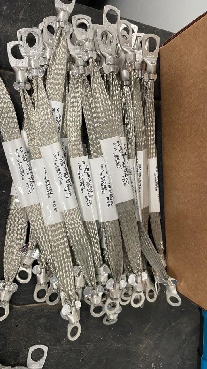 Braided silver grounding cables with ring terminals at both ends and white identification sleeves, arranged on a workbench.